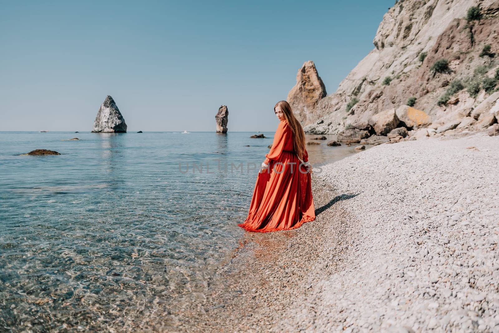 Woman travel sea. Happy tourist in red dress enjoy taking picture outdoors for memories. Woman traveler posing on the rock at sea bay surrounded by volcanic mountains, sharing travel adventure journey by panophotograph