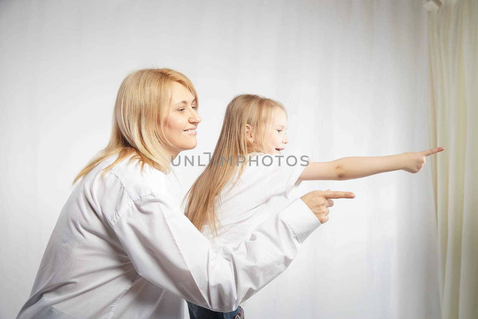 Portrait of a blonde mother and daughter who having communicate and play on white background. Mom and little girl models pointing his finger forward. The concept of family unity