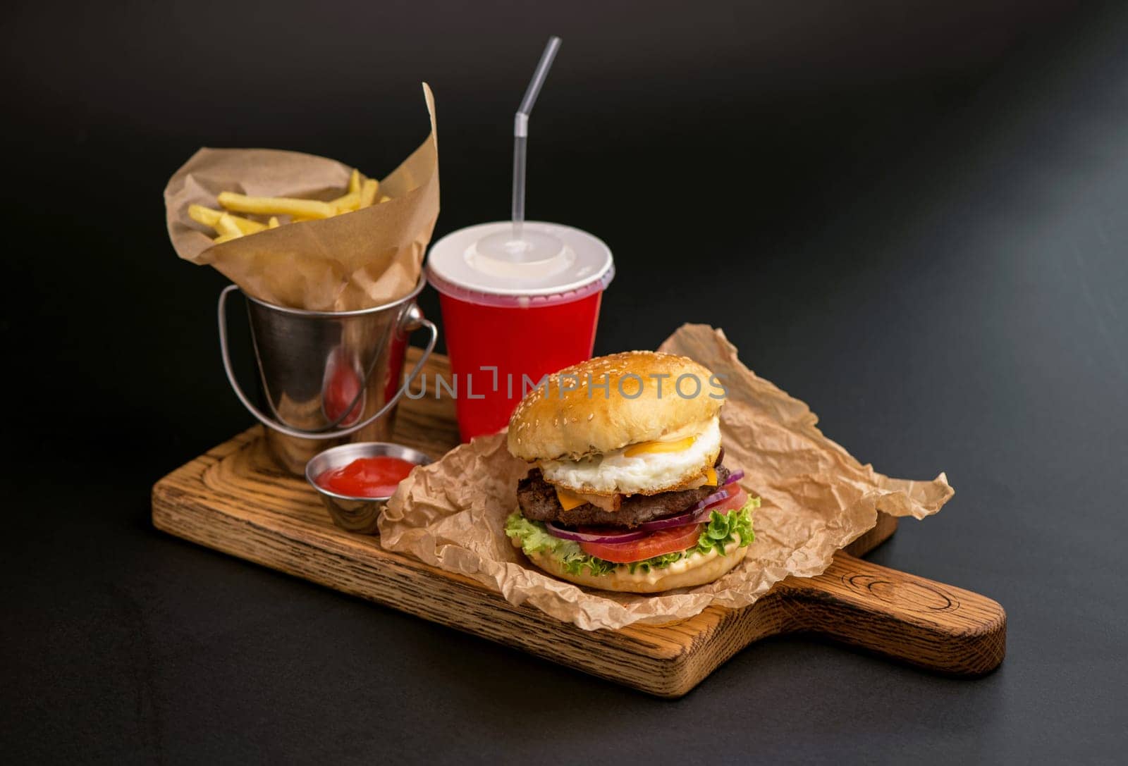 Classic burgers with becon, meat, cheese, onion, tomato and cheese and integral bun. On textured black board. Close up. Fried potatoes and glass of cola.