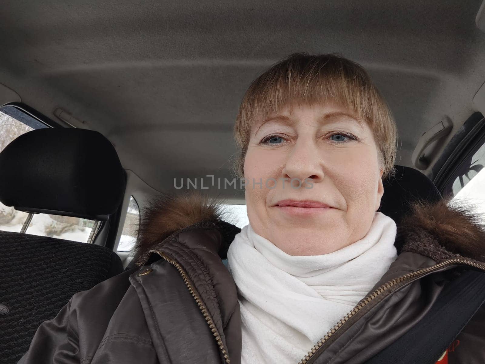 Portrait of adult cute funny blonde woman in warm clothes taking a selfie inside the car and driving it. The concept of a pleasant trip in spring, autumn, winter by keleny