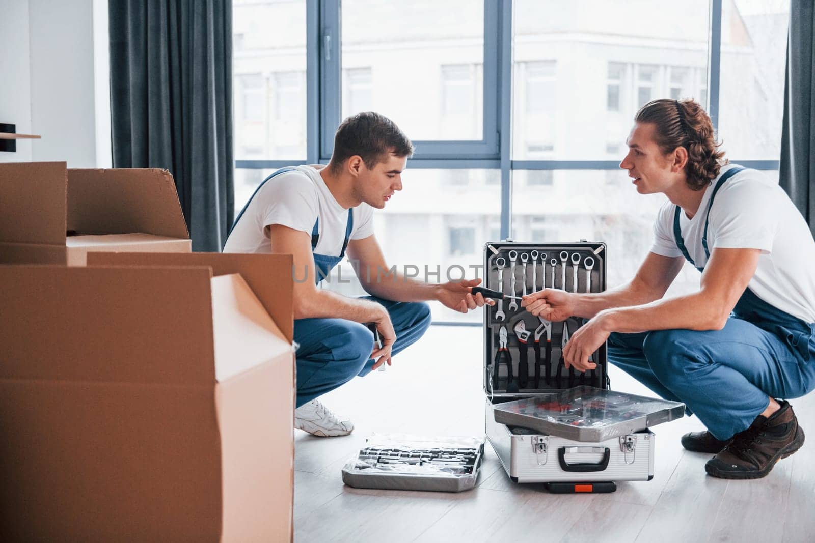 Uses tools from case. Two young movers in blue uniform working indoors in the room by Standret