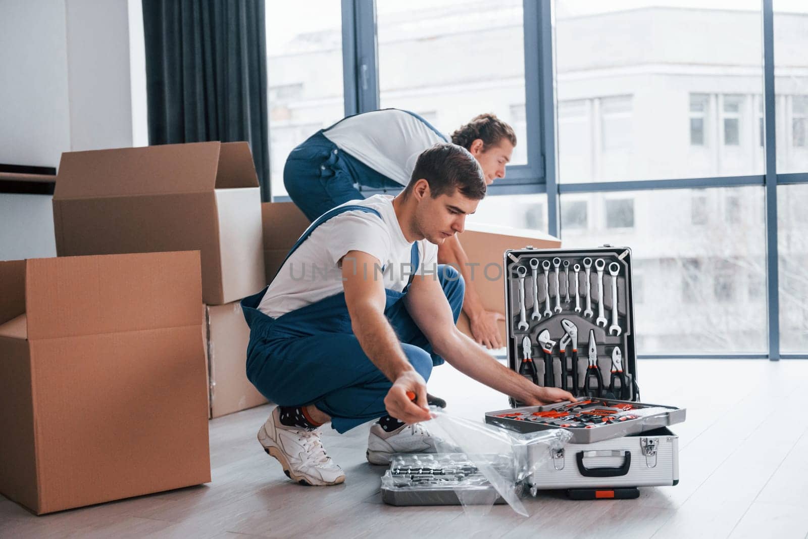 Uses tools from case. Two young movers in blue uniform working indoors in the room by Standret