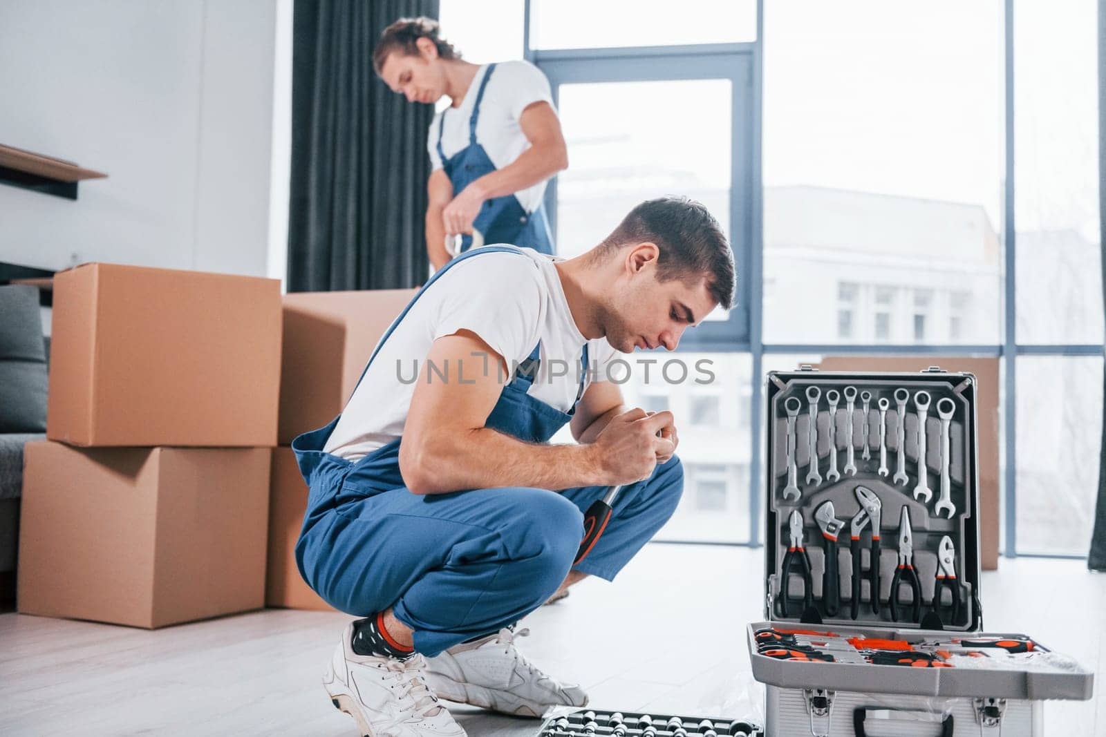 Uses tools from case. Two young movers in blue uniform working indoors in the room.