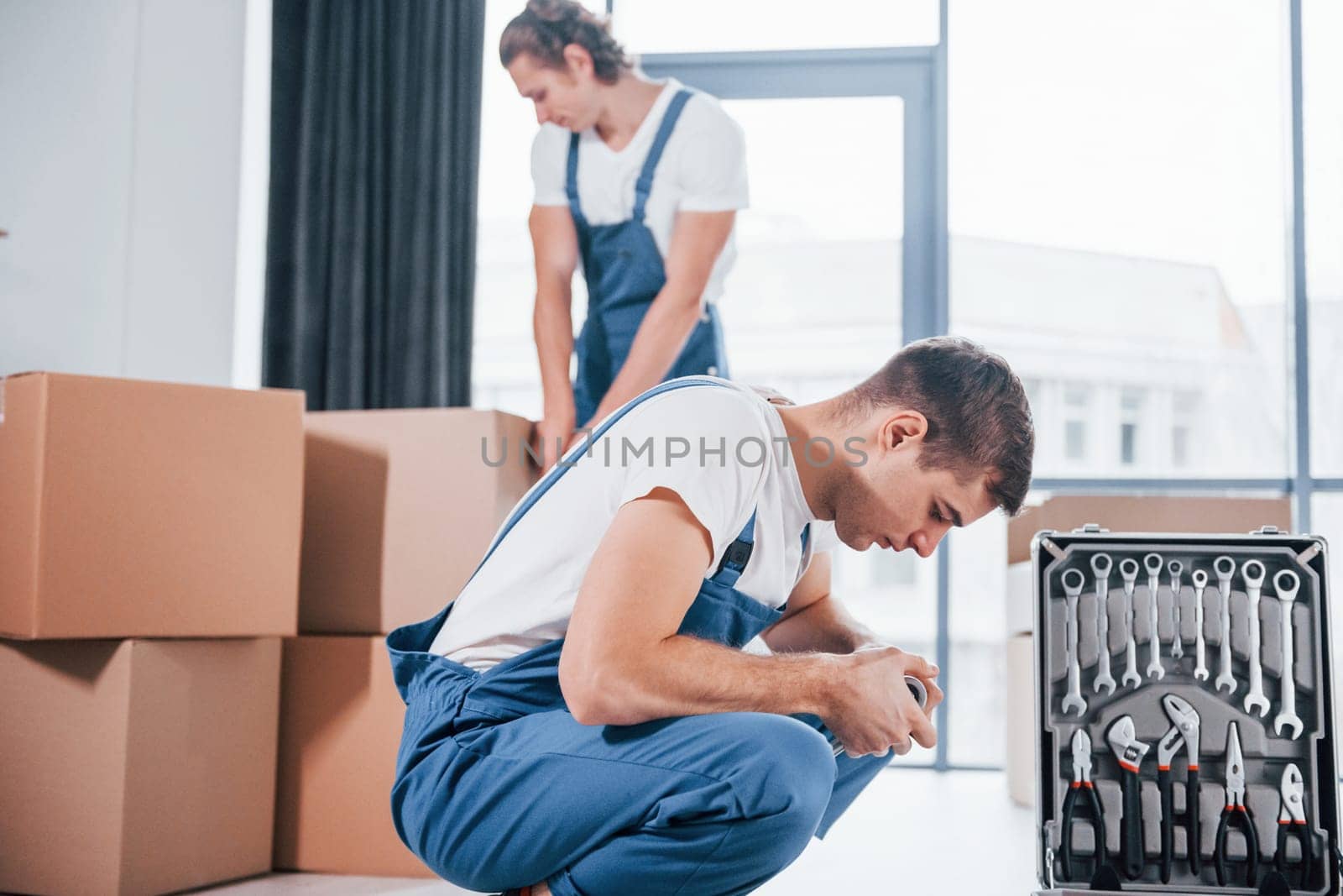 Uses tools from case. Two young movers in blue uniform working indoors in the room by Standret
