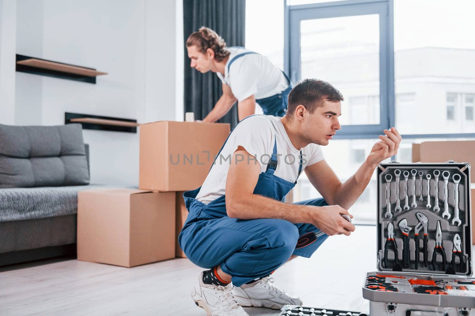 Uses tools from case. Two young movers in blue uniform working indoors in the room by Standret