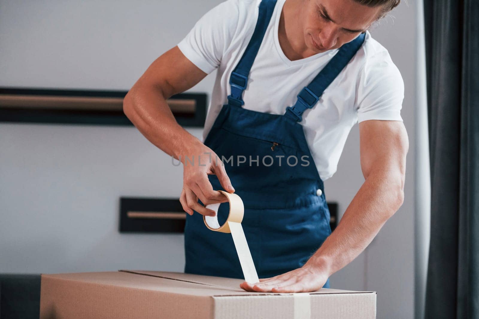 Packaging the box. Young male mover in blue uniform works indoors in the room.