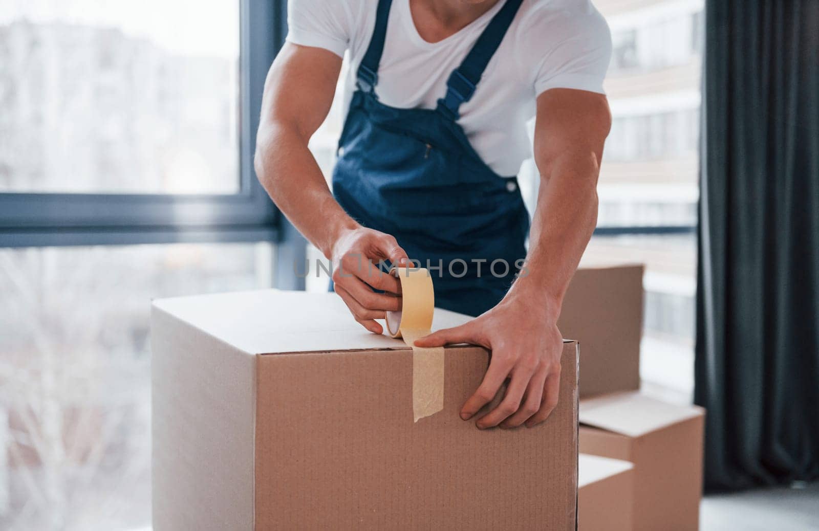 Packaging the box. Young male mover in blue uniform works indoors in the room.
