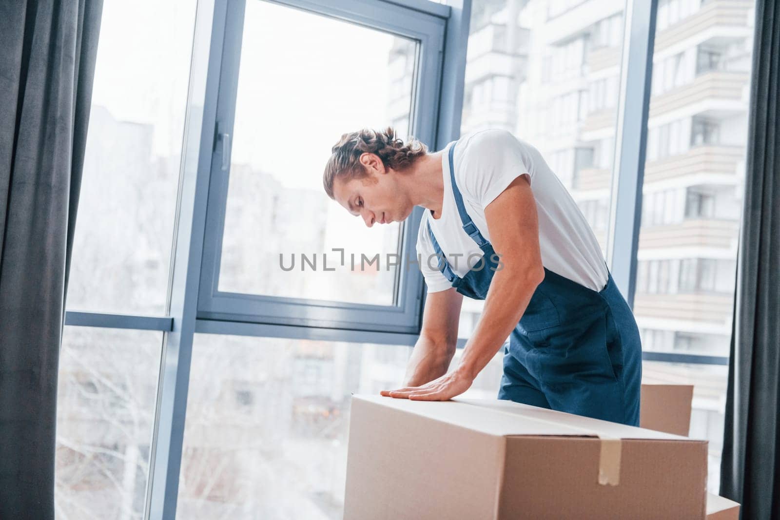 Packaging the box. Young male mover in blue uniform works indoors in the room.