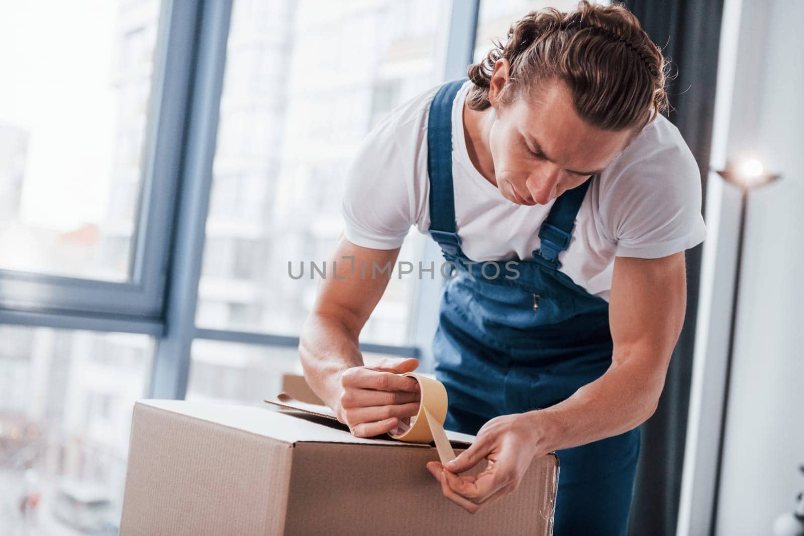 Packaging the box. Young male mover in blue uniform works indoors in the room.