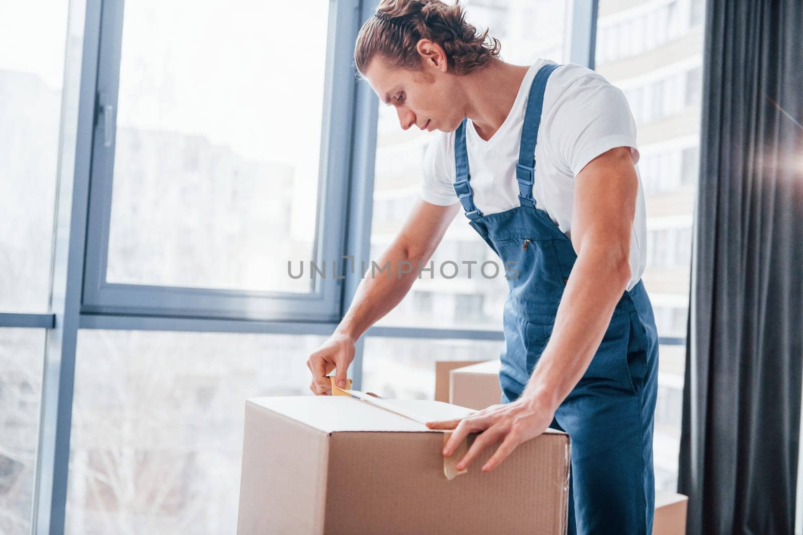 Packaging the box. Young male mover in blue uniform works indoors in the room.