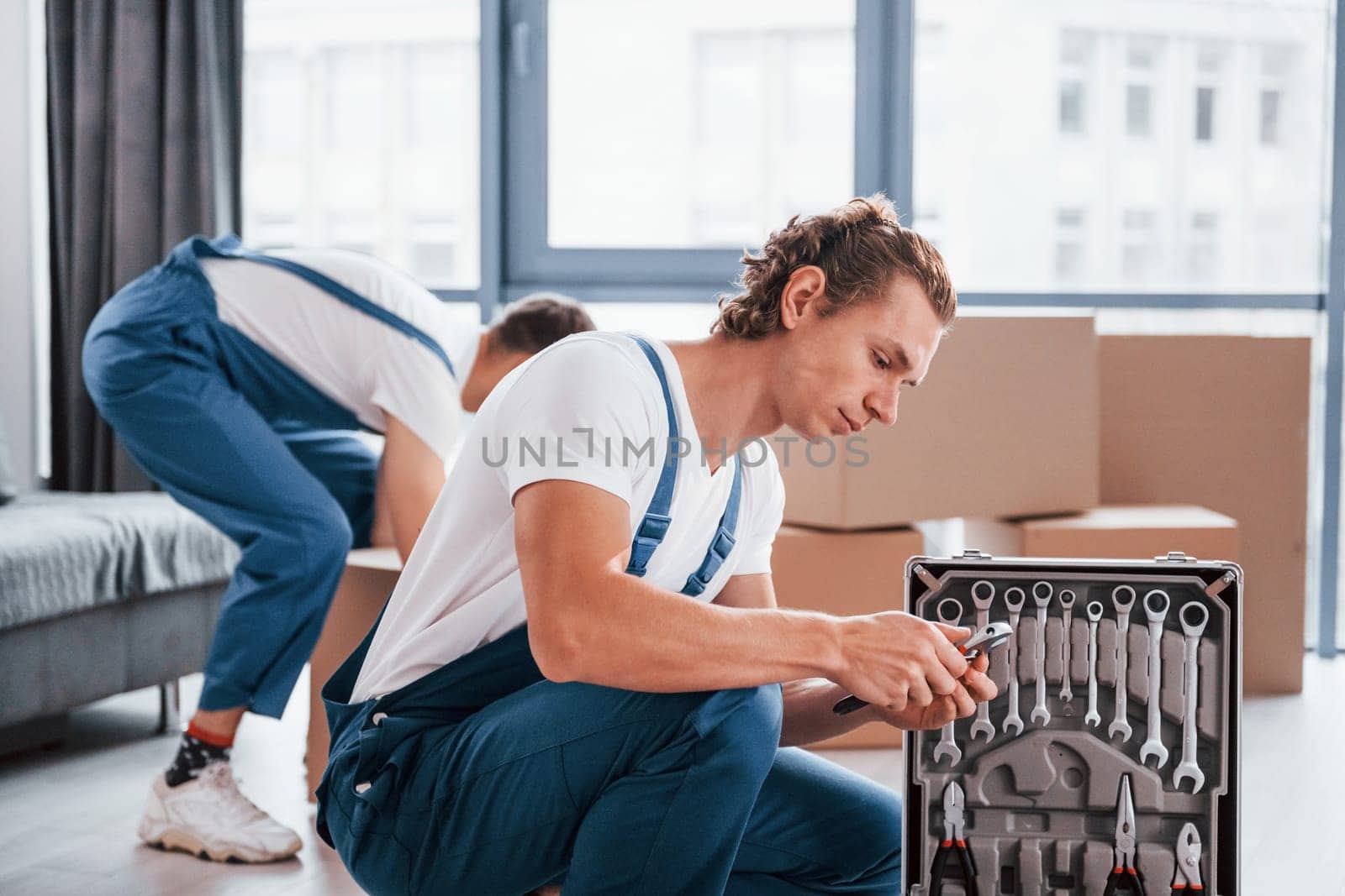 Uses tools from case. Two young movers in blue uniform working indoors in the room.