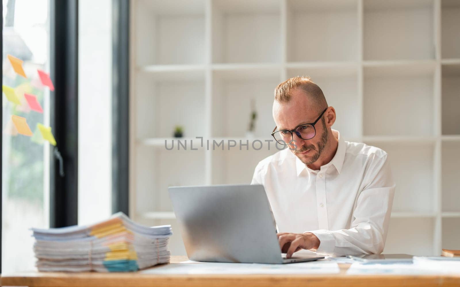 Young business man wearing glasses using laptop, typing on keyboard, writing email or message, sitting in office.