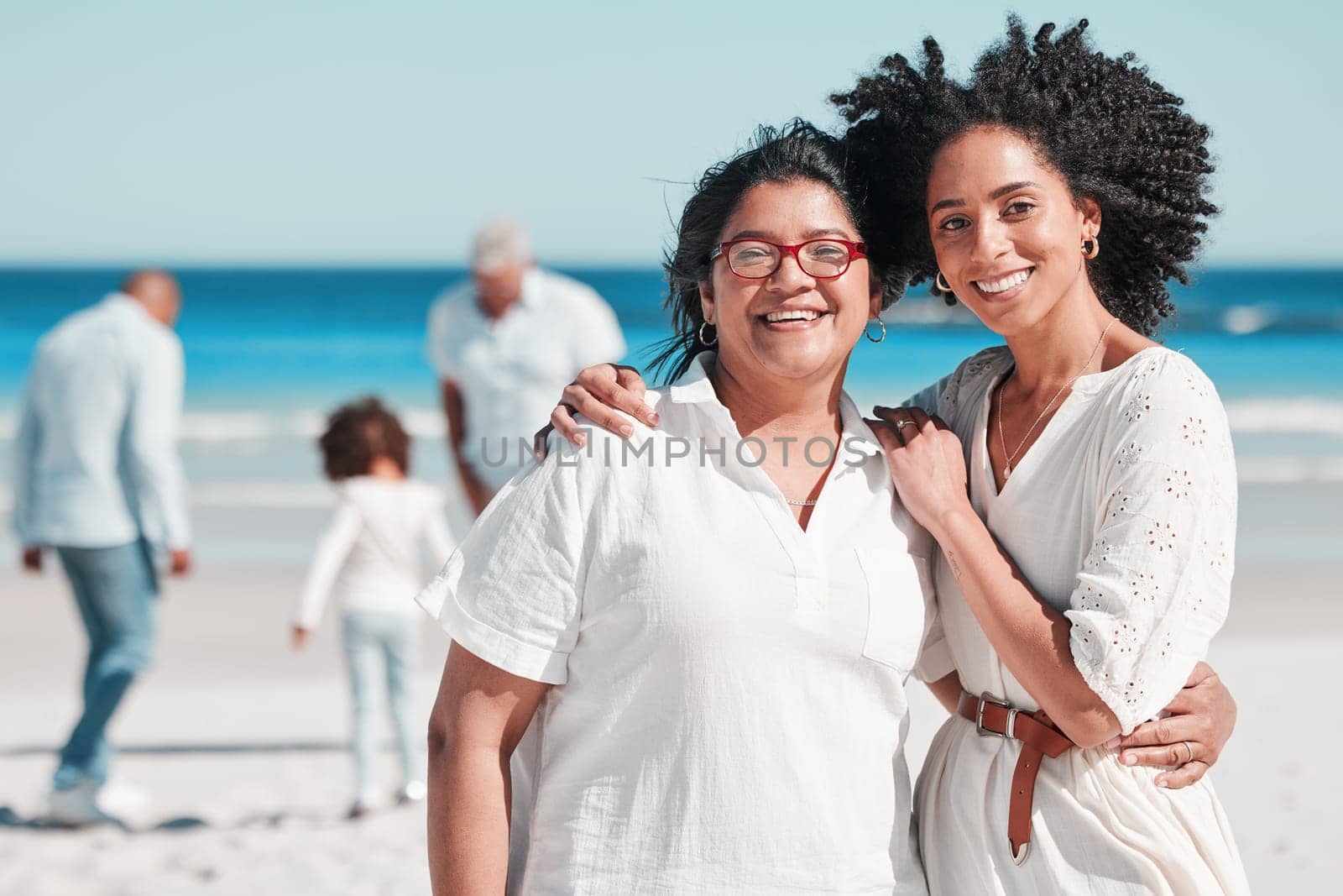 Portrait, nature with a mother and daughter on the beach during summer while their family play in the background. Love, smile or summer with a senior woman and adult child bonding outdoor together.