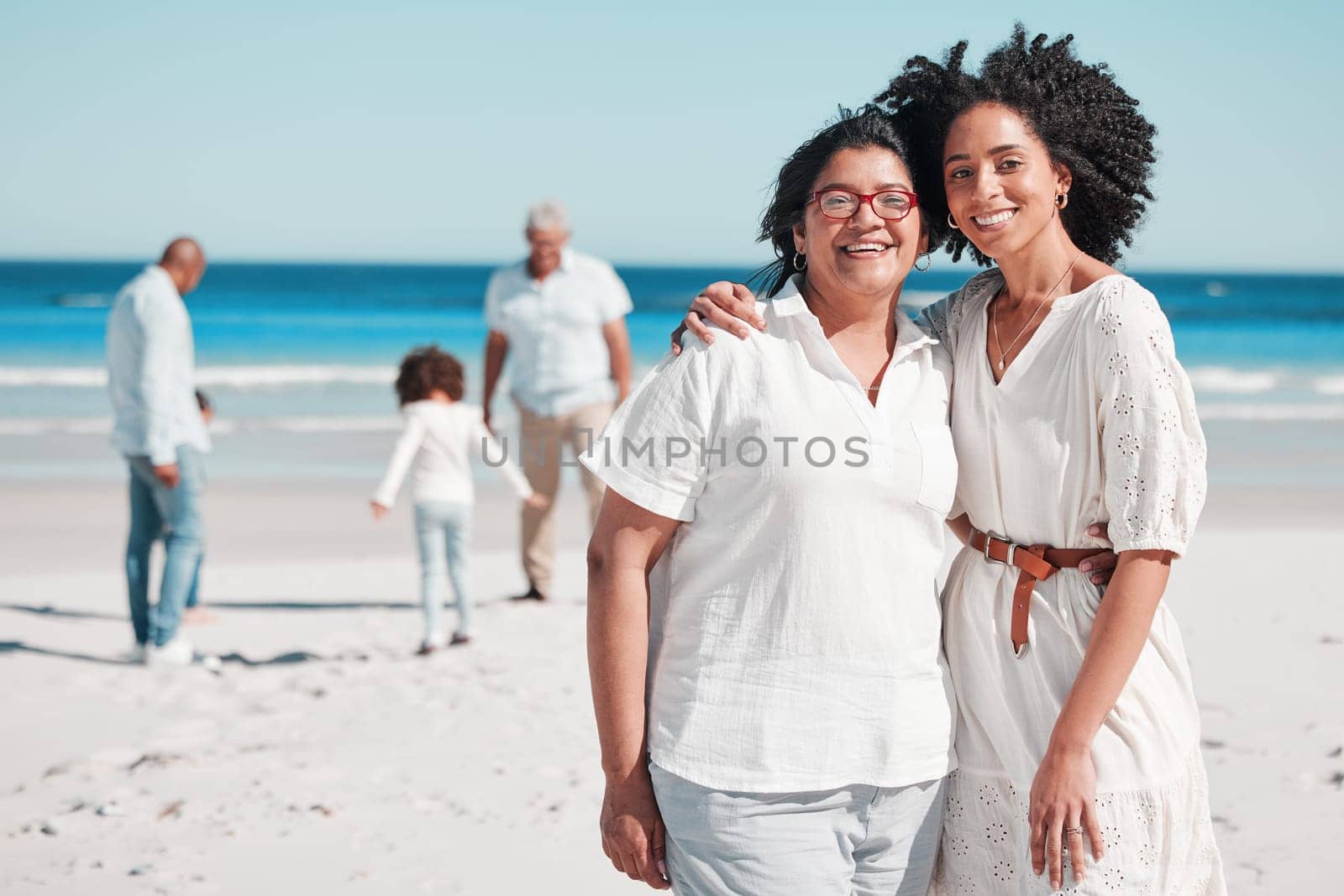 Portrait, summer with a mother and daughter on the beach during summer while their family play in the background. Love, smile or nature with a senior woman and adult child bonding outdoor together.