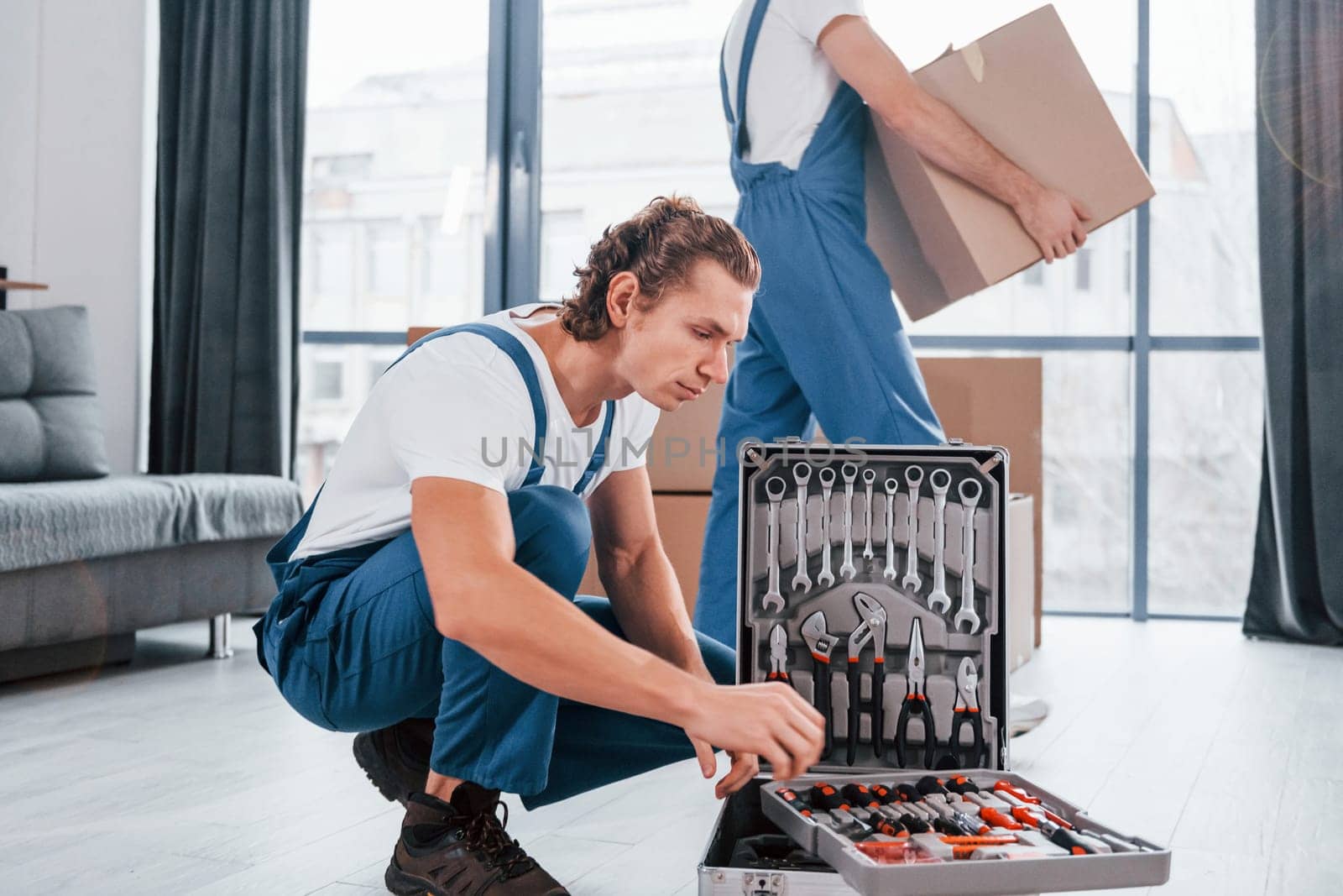 Uses tools from case. Two young movers in blue uniform working indoors in the room by Standret