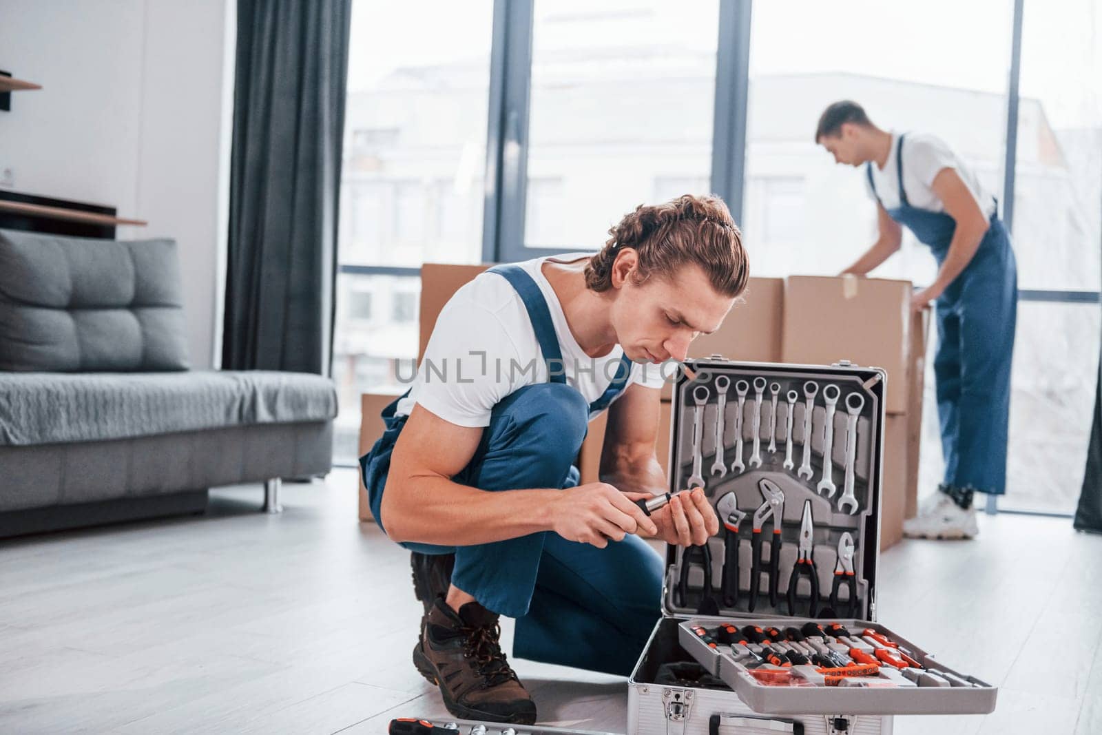 Case with equipment. Two young movers in blue uniform working indoors in the room by Standret