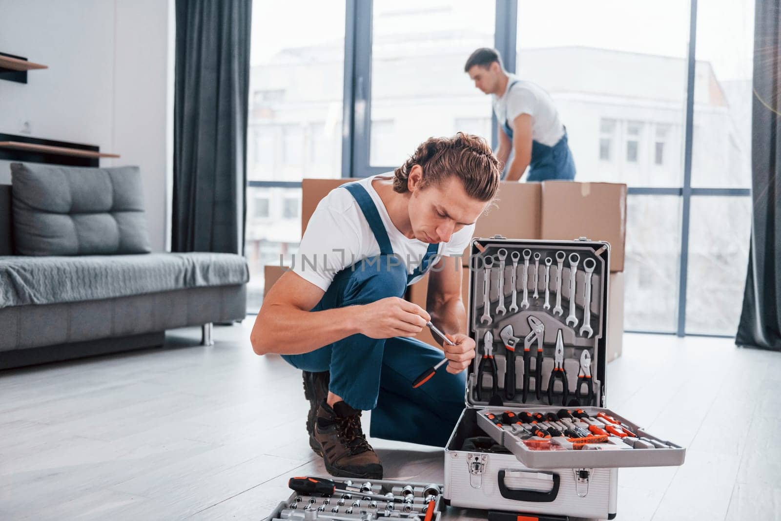 Case with equipment. Two young movers in blue uniform working indoors in the room by Standret