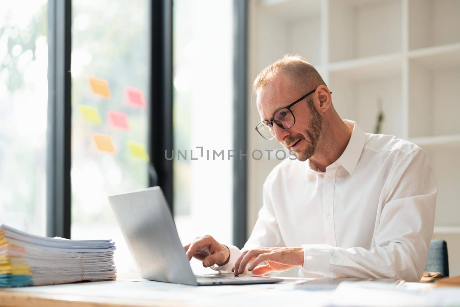 Young business man wearing glasses using laptop, typing on keyboard, writing email or message, sitting in office by nateemee