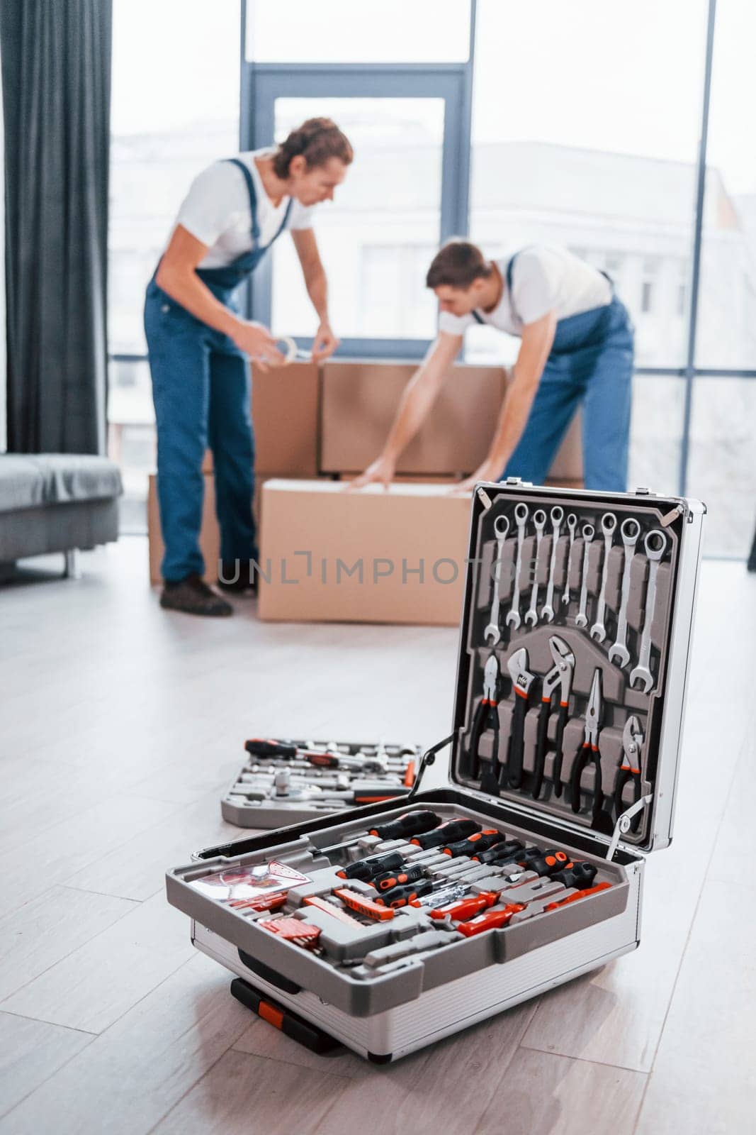 Case with equipment. Two young movers in blue uniform working indoors in the room by Standret