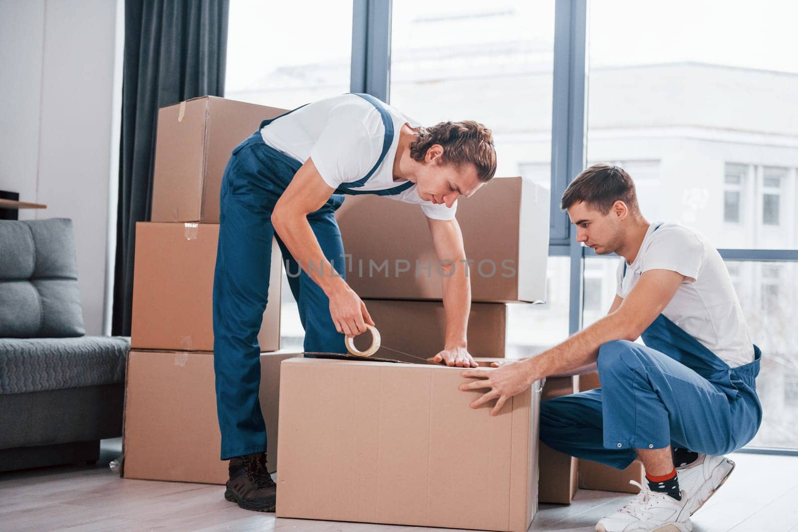 Packaging the box. Two young movers in blue uniform working indoors in the room by Standret