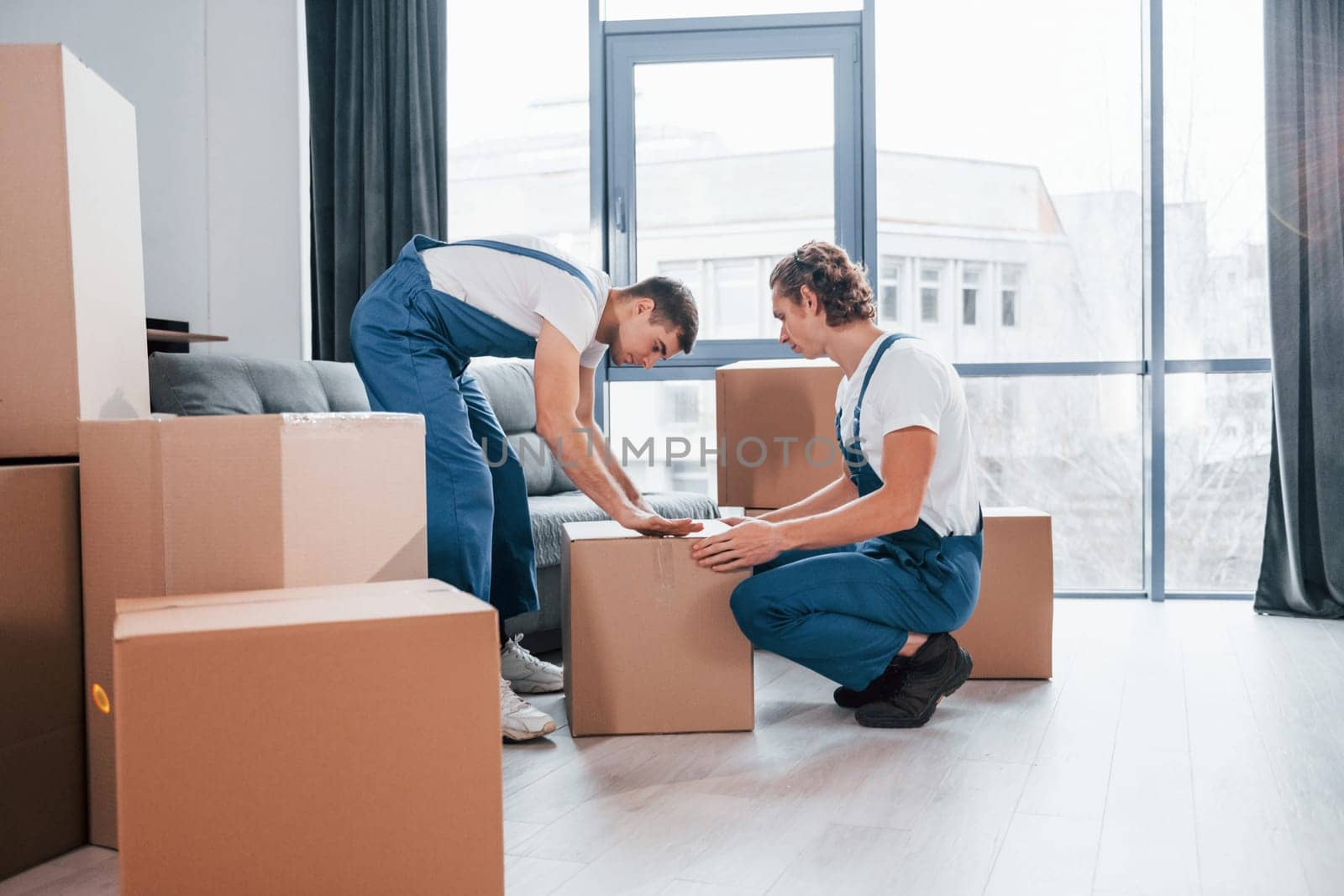 Packaging the box. Two young movers in blue uniform working indoors in the room.