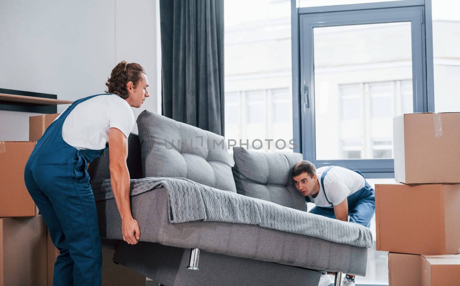 Carrying heavy sofa. Two young movers in blue uniform working indoors in the room.