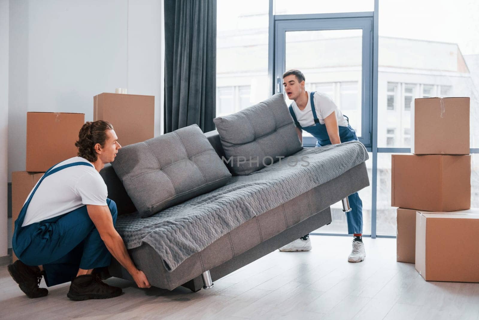 Carrying heavy sofa. Two young movers in blue uniform working indoors in the room by Standret
