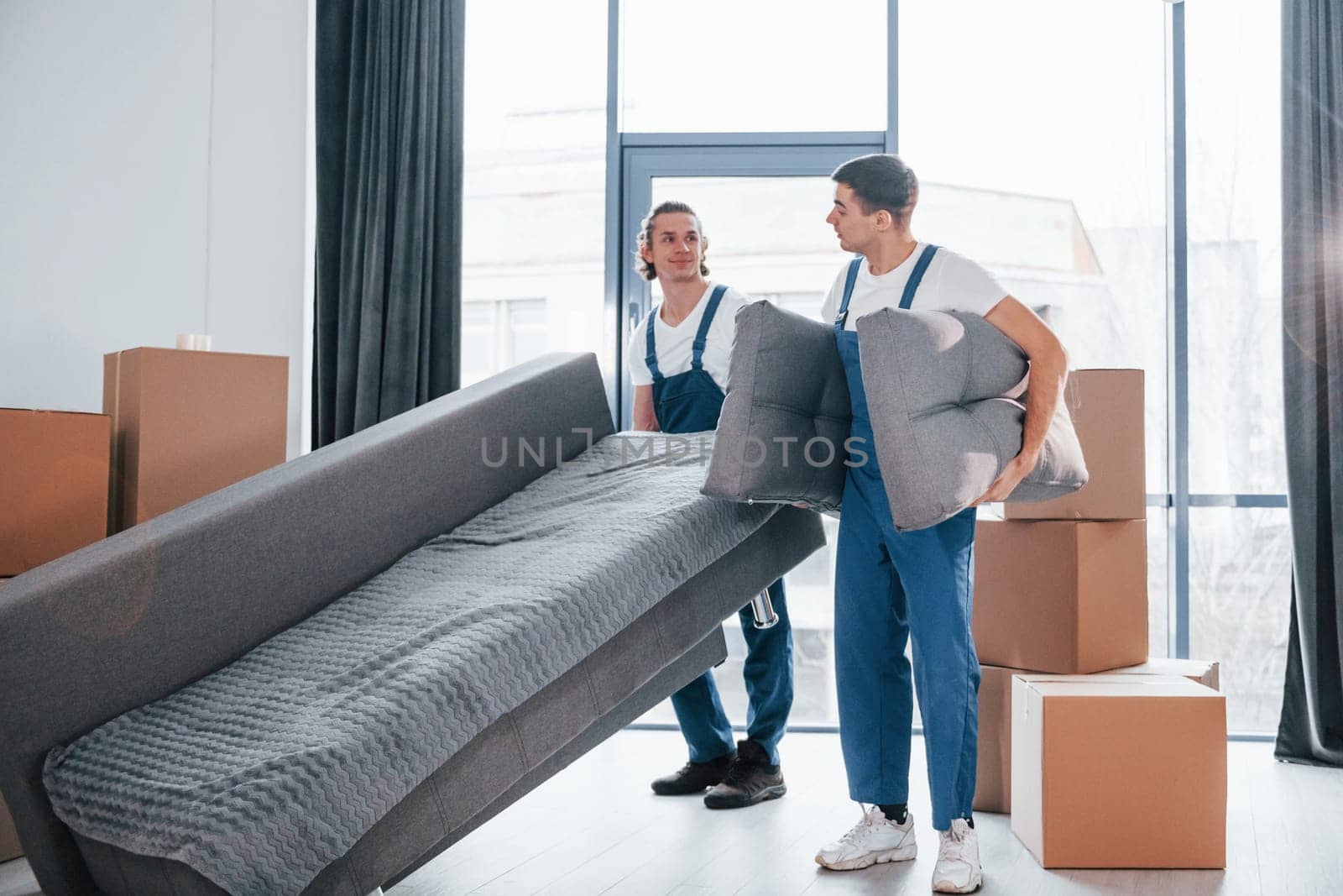 Carrying heavy sofa. Two young movers in blue uniform working indoors in the room by Standret