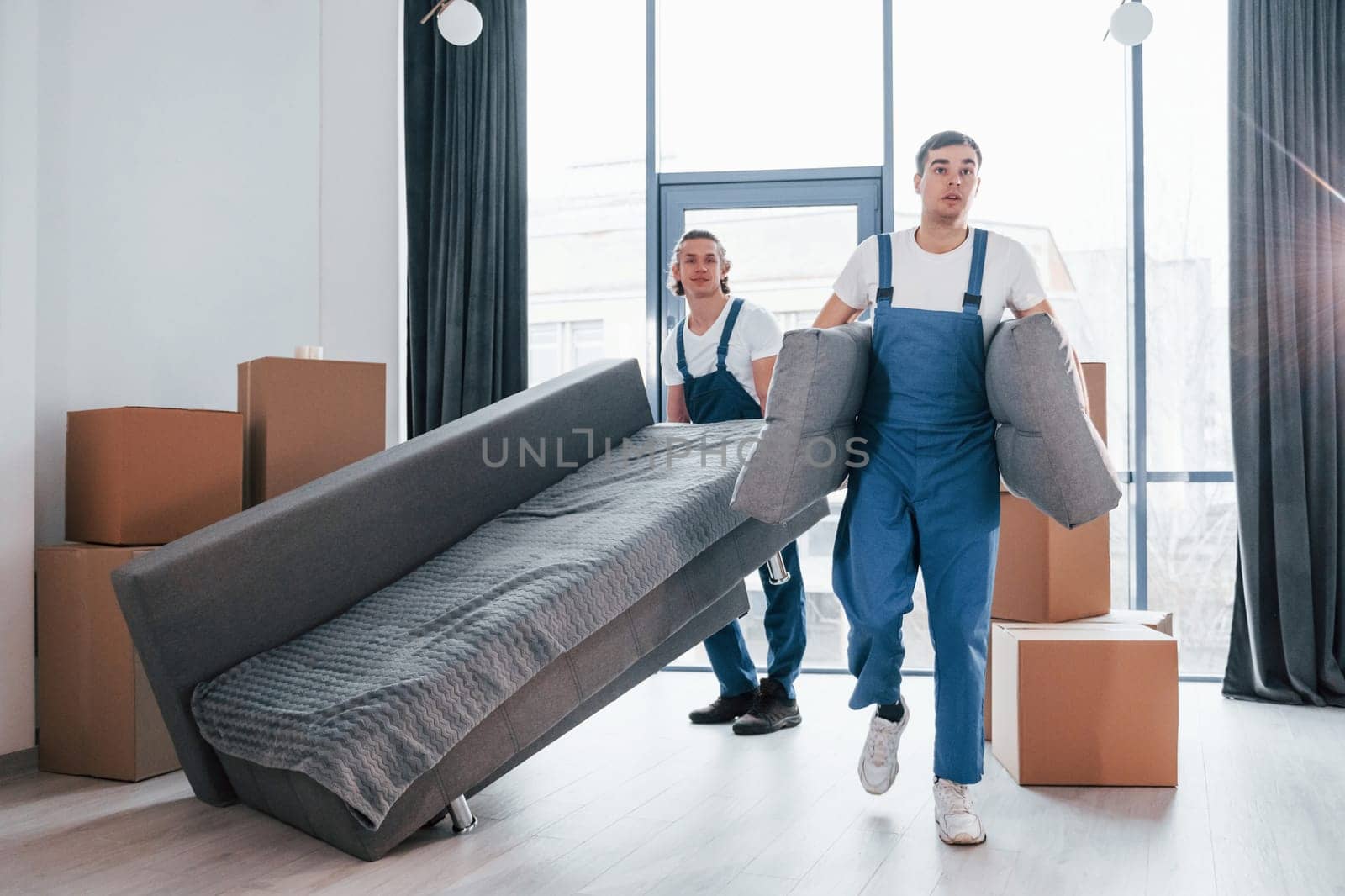 Carrying heavy sofa. Two young movers in blue uniform working indoors in the room by Standret