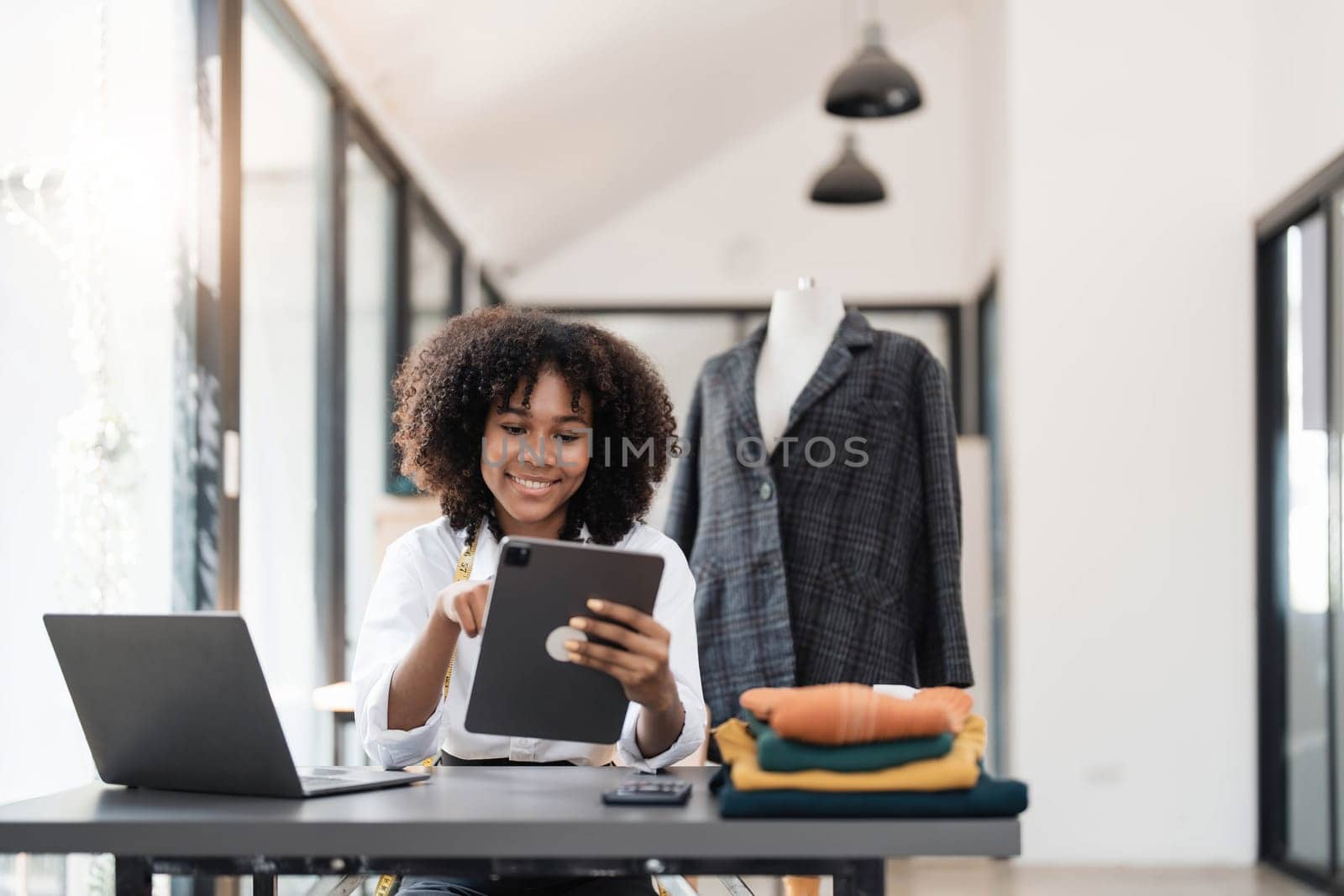 Portrait of young female tailor with tape measure in her neck sitting in stylish dress and holding digital tablet in office by wichayada