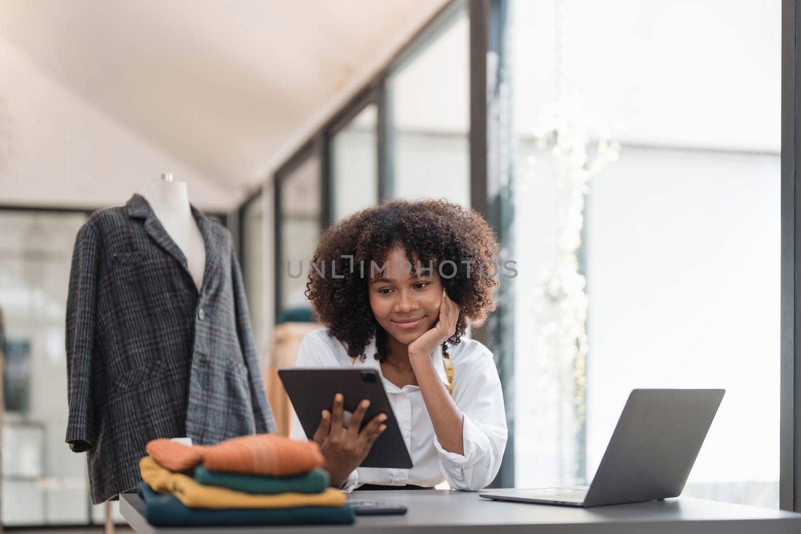 Portrait of young female tailor with tape measure in her neck sitting in stylish dress and holding digital tablet in office by wichayada