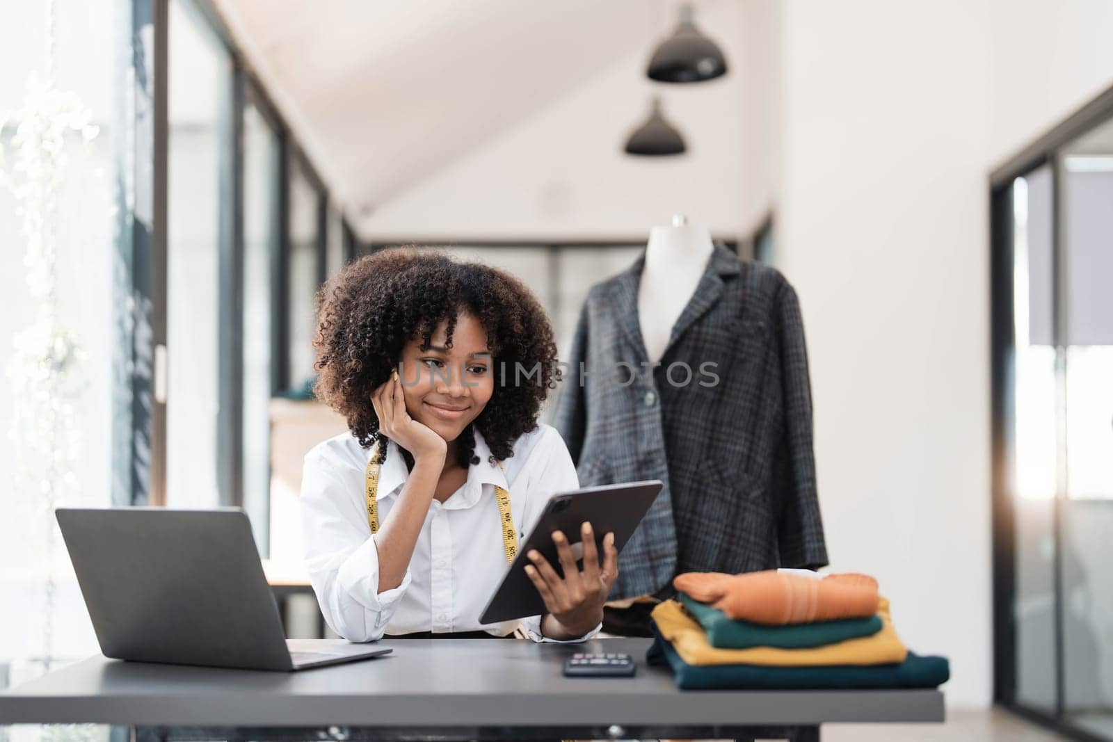 Portrait of young female tailor with tape measure in her neck sitting in stylish dress and holding digital tablet in office by wichayada