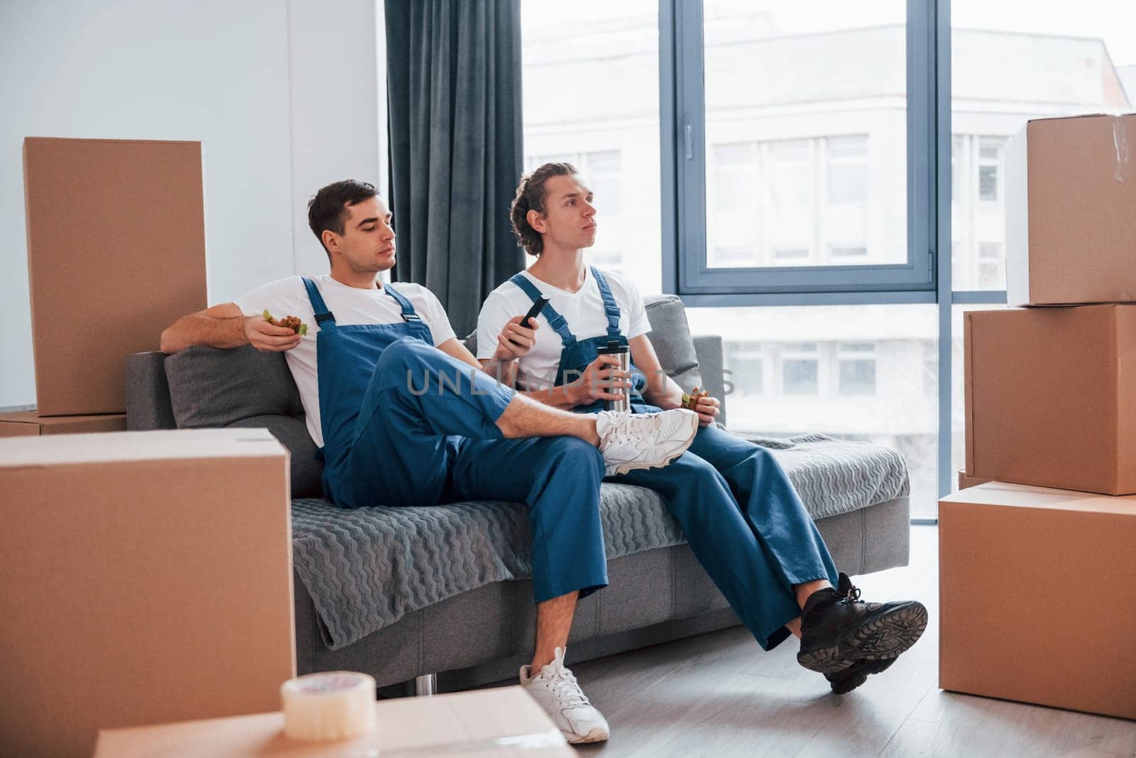 Sitting on the sofa. Tired and taking break. Two young movers in blue uniform working indoors in the room.