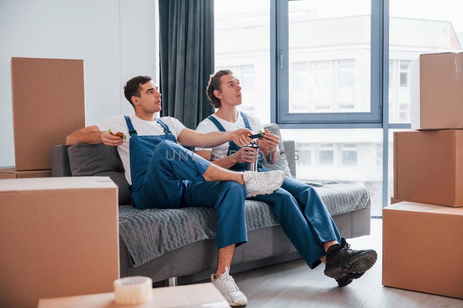 Sitting on the sofa. Tired and taking break. Two young movers in blue uniform working indoors in the room.