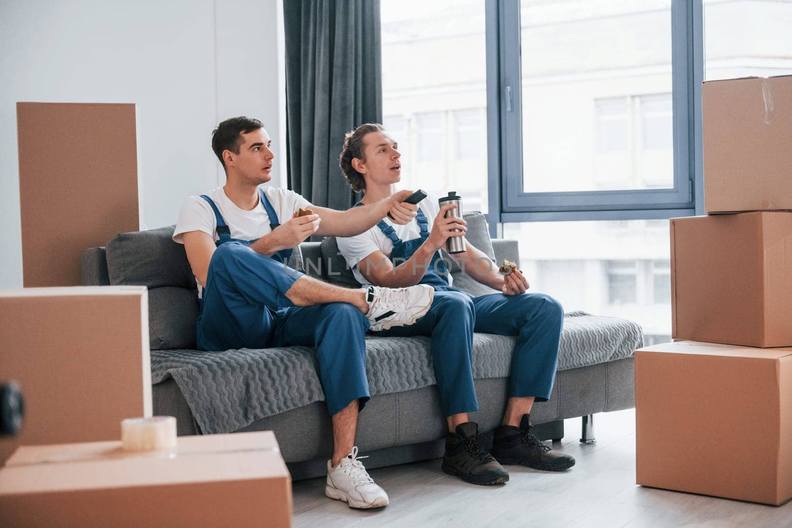 Sitting on the sofa. Tired and taking break. Two young movers in blue uniform working indoors in the room.