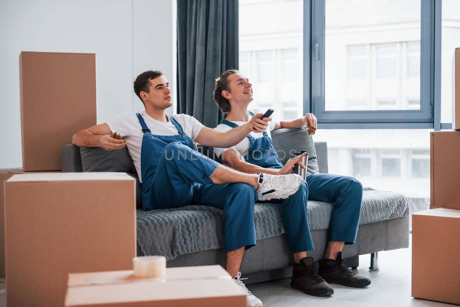 Sitting on the sofa. Tired and taking break. Two young movers in blue uniform working indoors in the room.