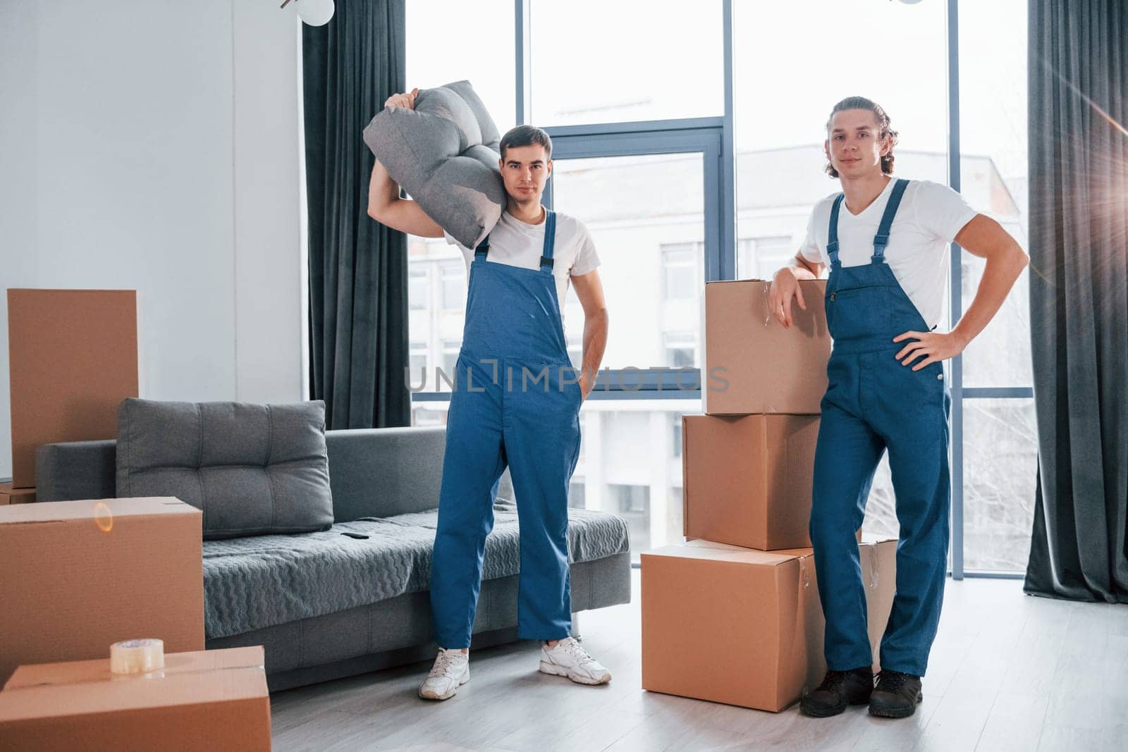 Busy day. Two young movers in blue uniform working indoors in the room.
