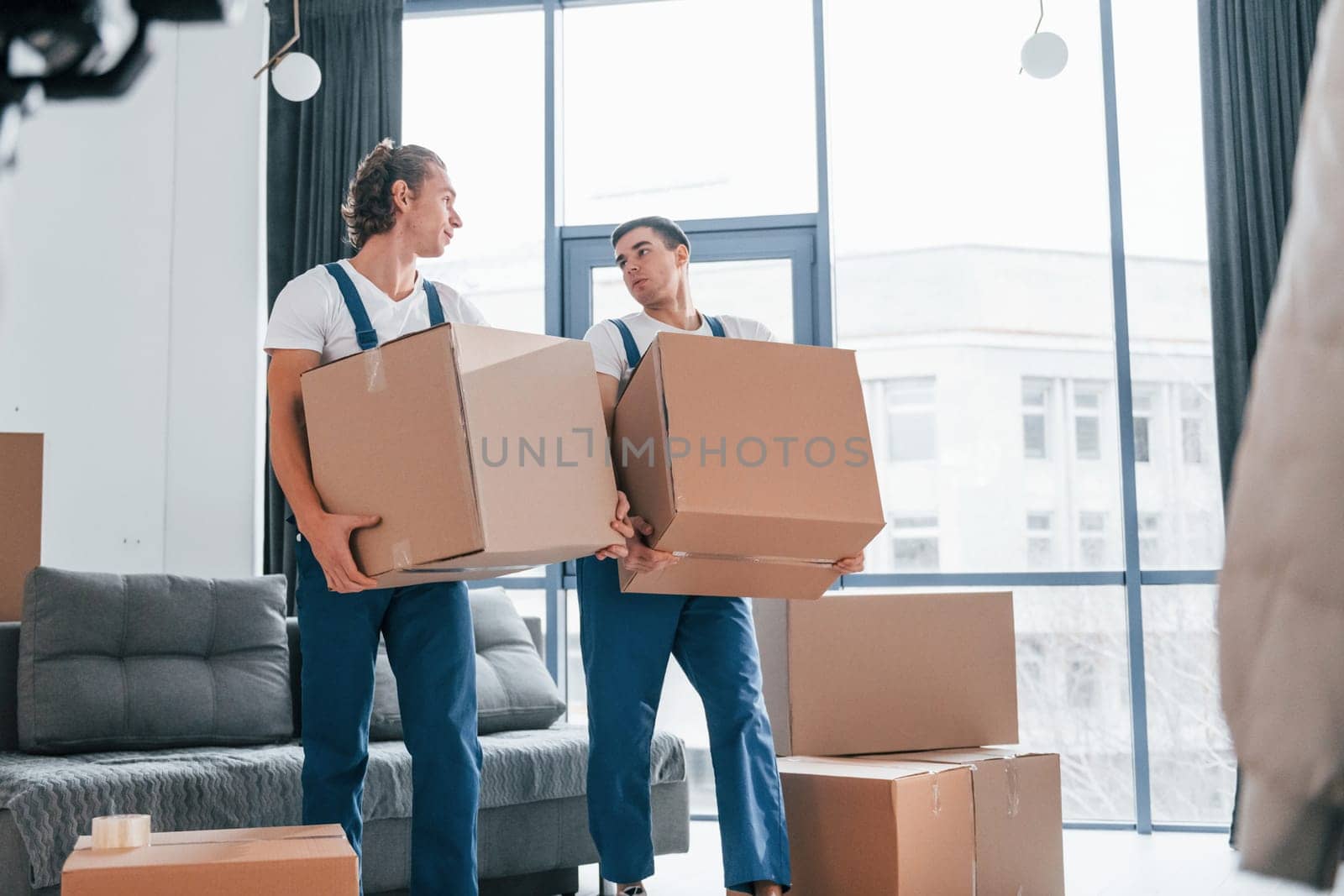 Busy day. Two young movers in blue uniform working indoors in the room by Standret