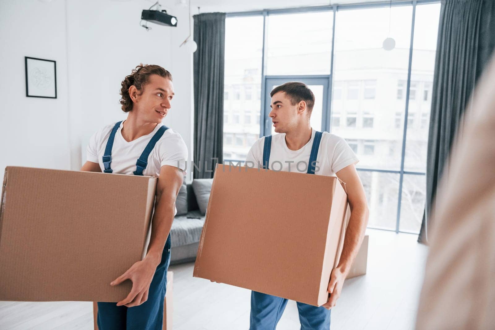 Busy day. Two young movers in blue uniform working indoors in the room.