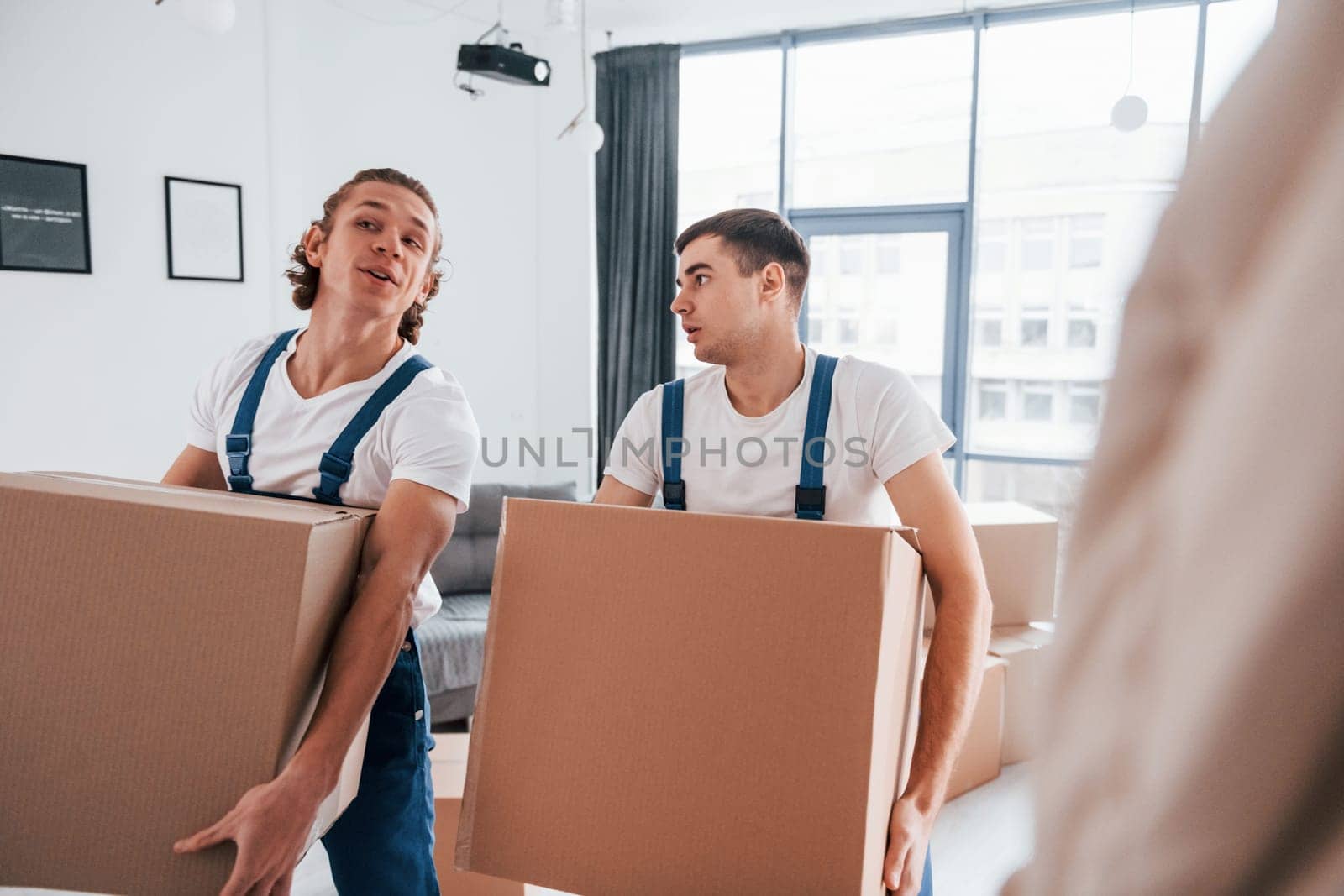 Busy day. Two young movers in blue uniform working indoors in the room.