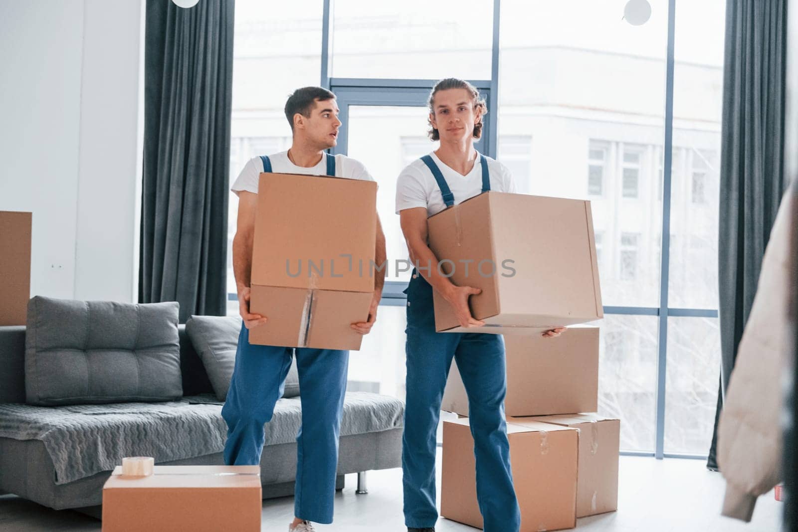 Busy day. Two young movers in blue uniform working indoors in the room.