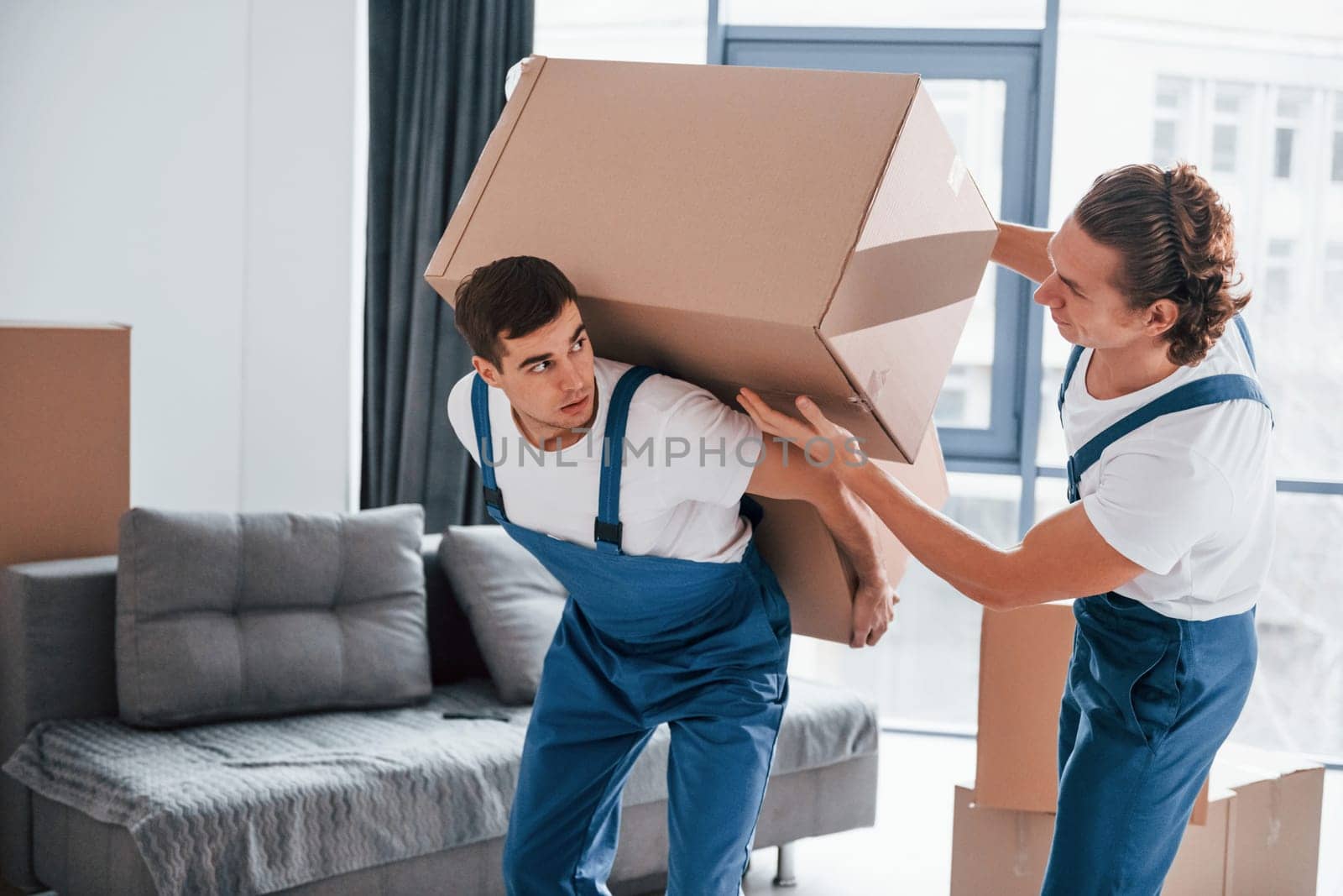 Heavy boxes. Two young movers in blue uniform working indoors in the room.