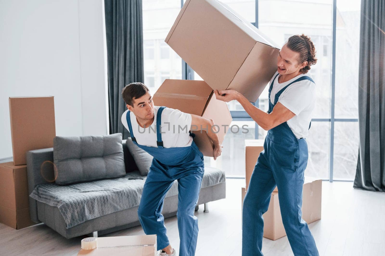 Heavy boxes. Two young movers in blue uniform working indoors in the room by Standret