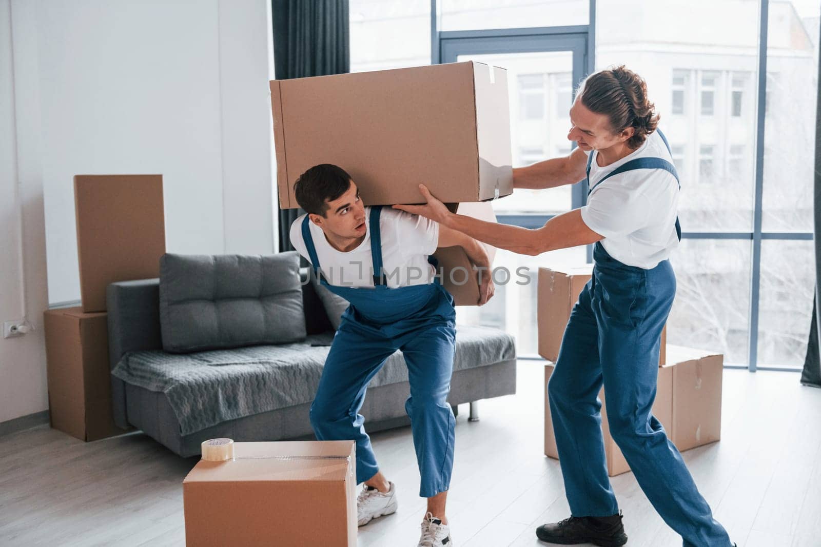 Heavy boxes. Two young movers in blue uniform working indoors in the room.