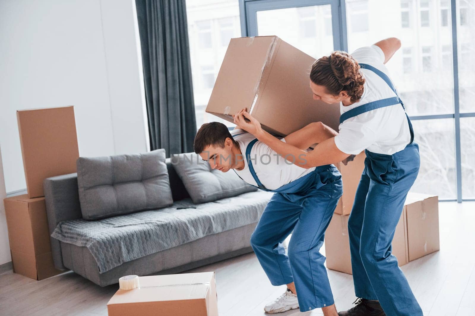 Heavy boxes. Two young movers in blue uniform working indoors in the room.