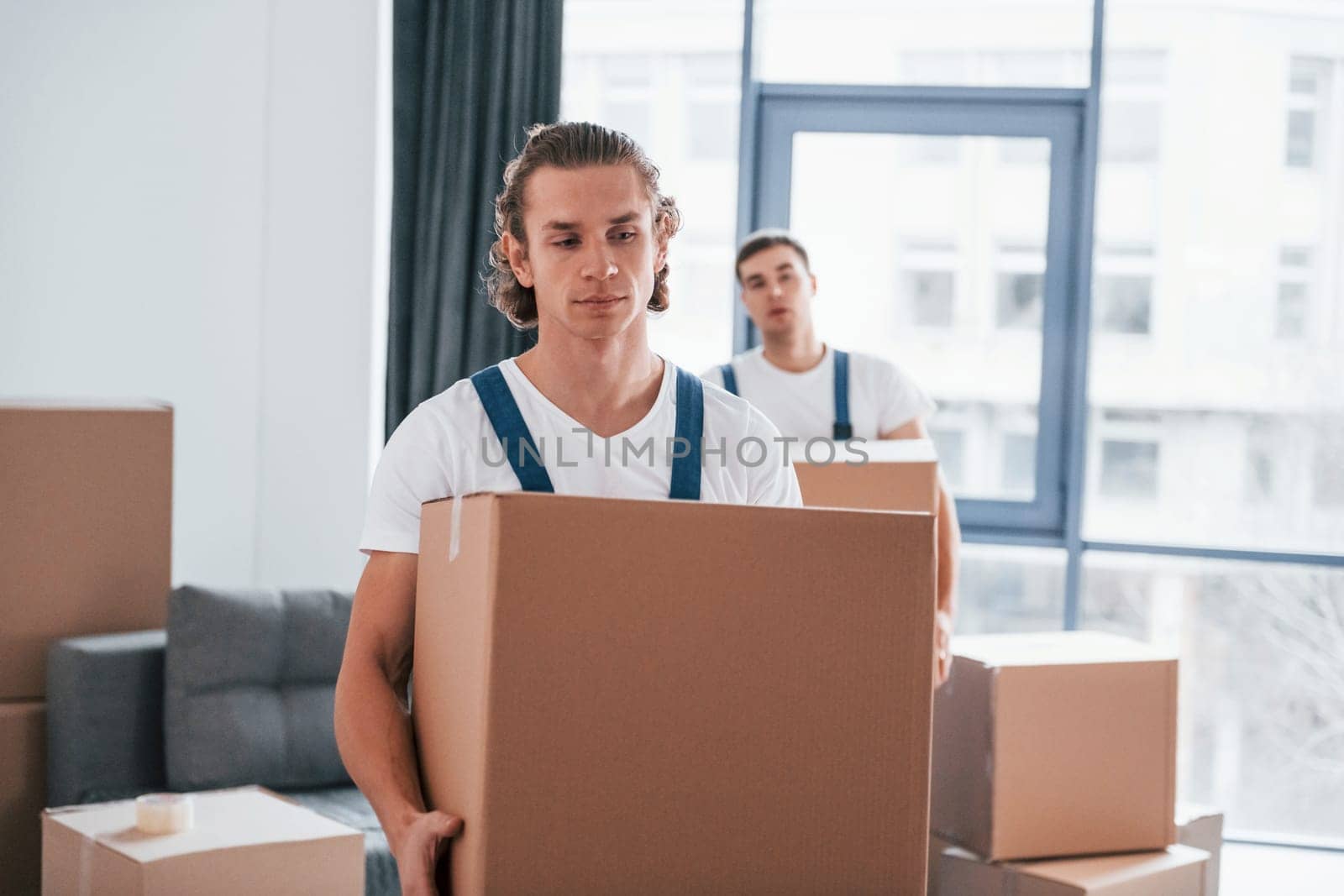 With boxes in hands. Two young movers in blue uniform working indoors in the room.