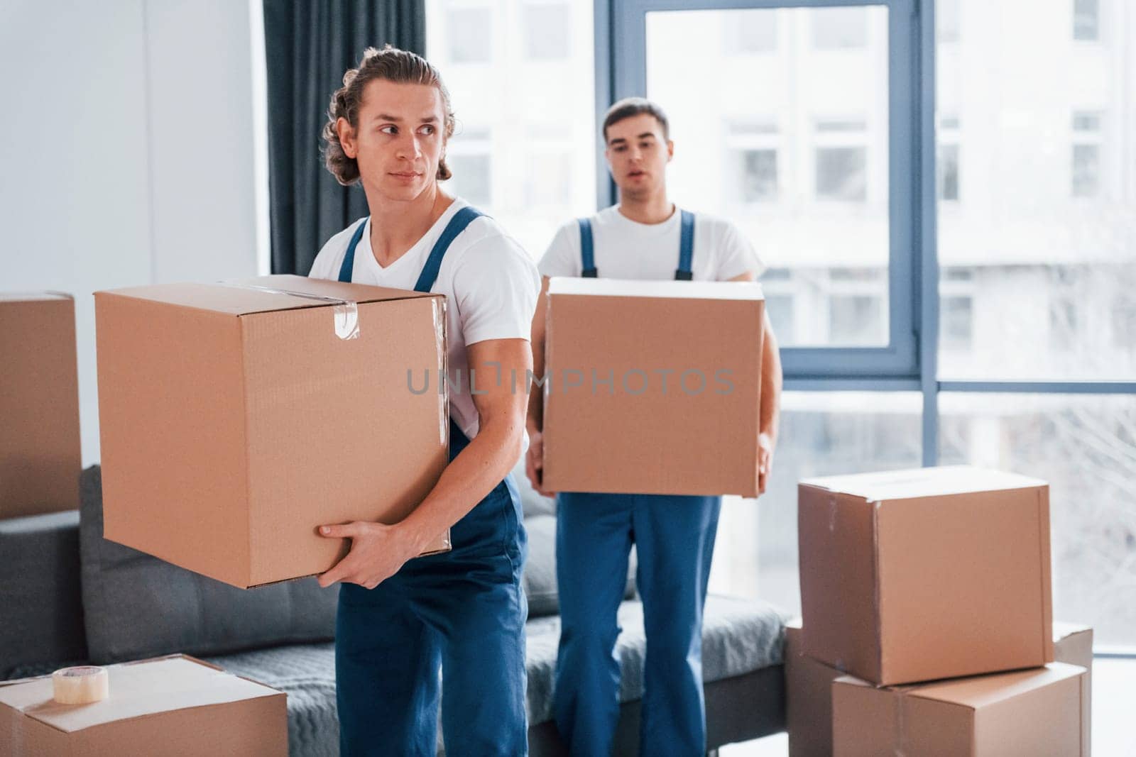 With boxes in hands. Two young movers in blue uniform working indoors in the room.