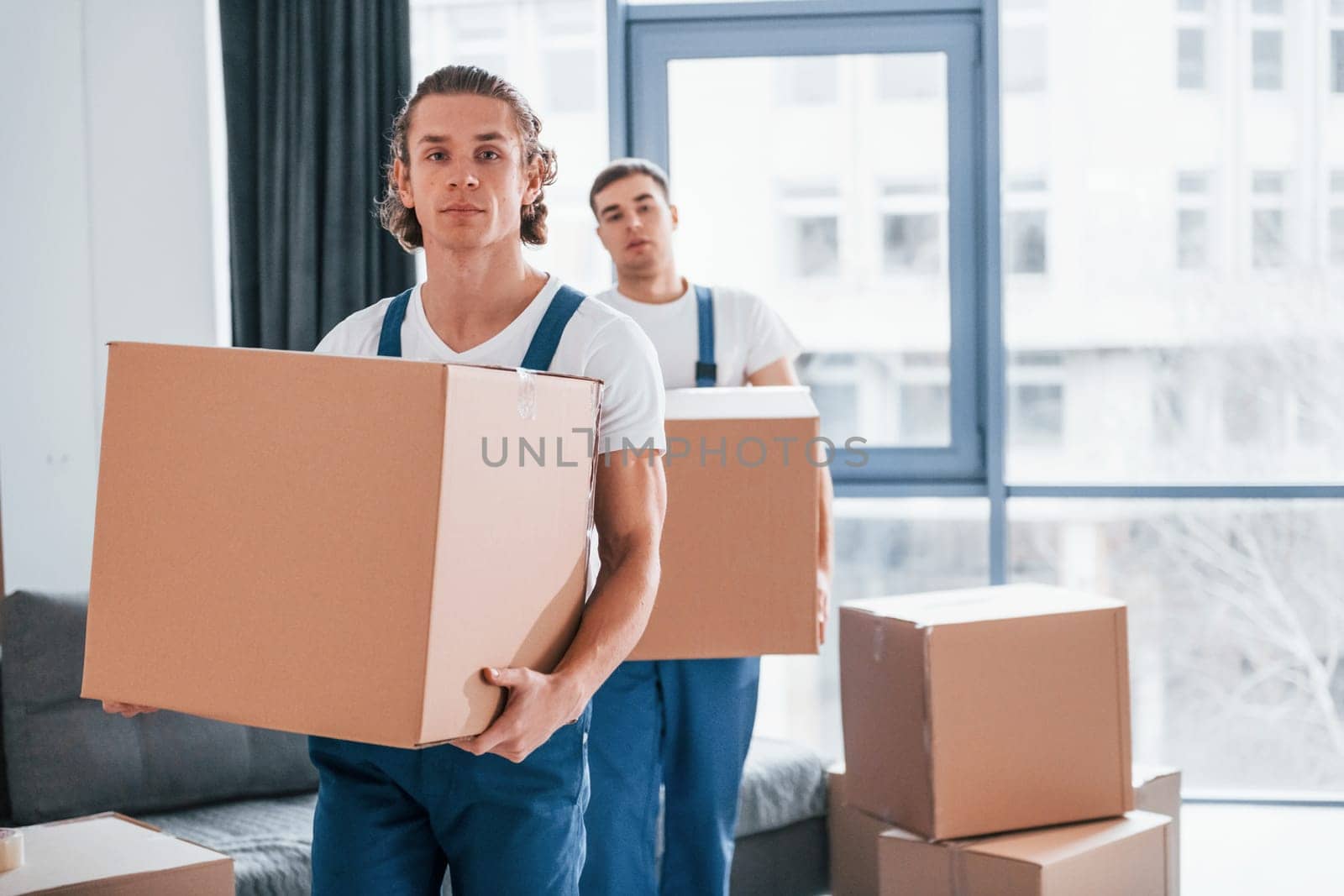 With boxes in hands. Two young movers in blue uniform working indoors in the room.
