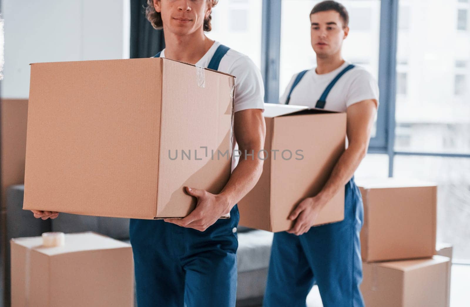 With boxes in hands. Two young movers in blue uniform working indoors in the room.