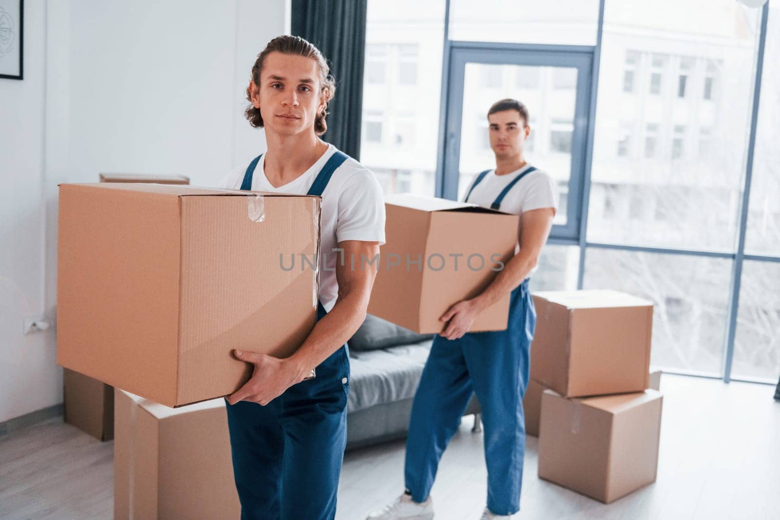 With boxes in hands. Two young movers in blue uniform working indoors in the room.